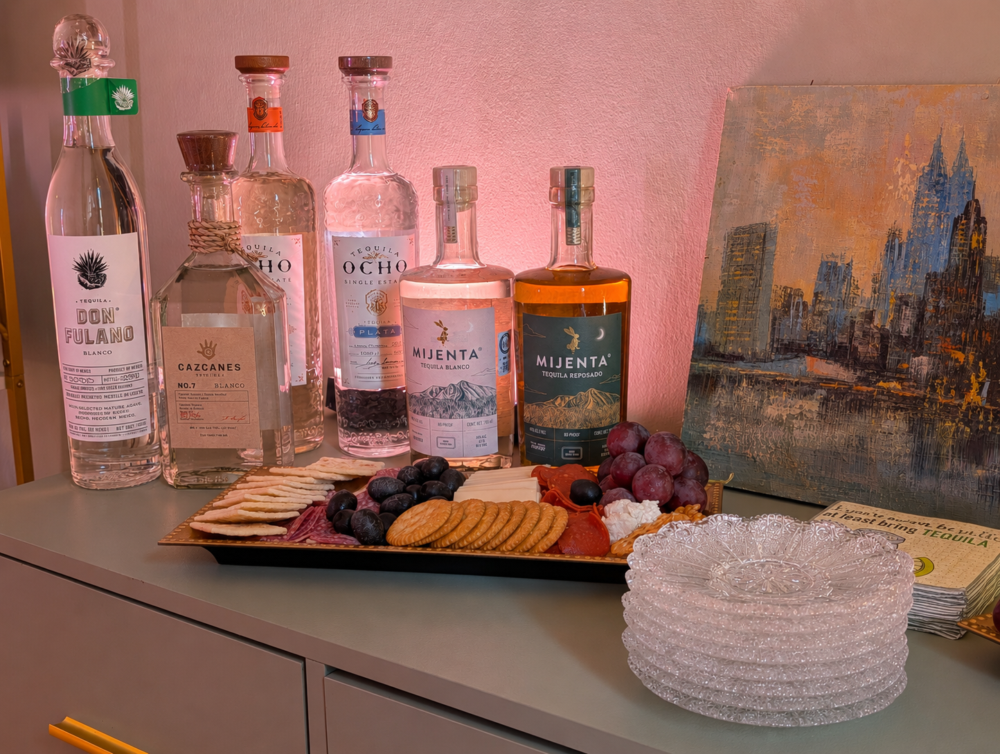 Assorted bottles of tequila, a cheese and cracker platter with grapes, cheese, meats, and crackers, a stack of clear glass plates, and a cityscape painting on a pink wall, all on a light-colored table.