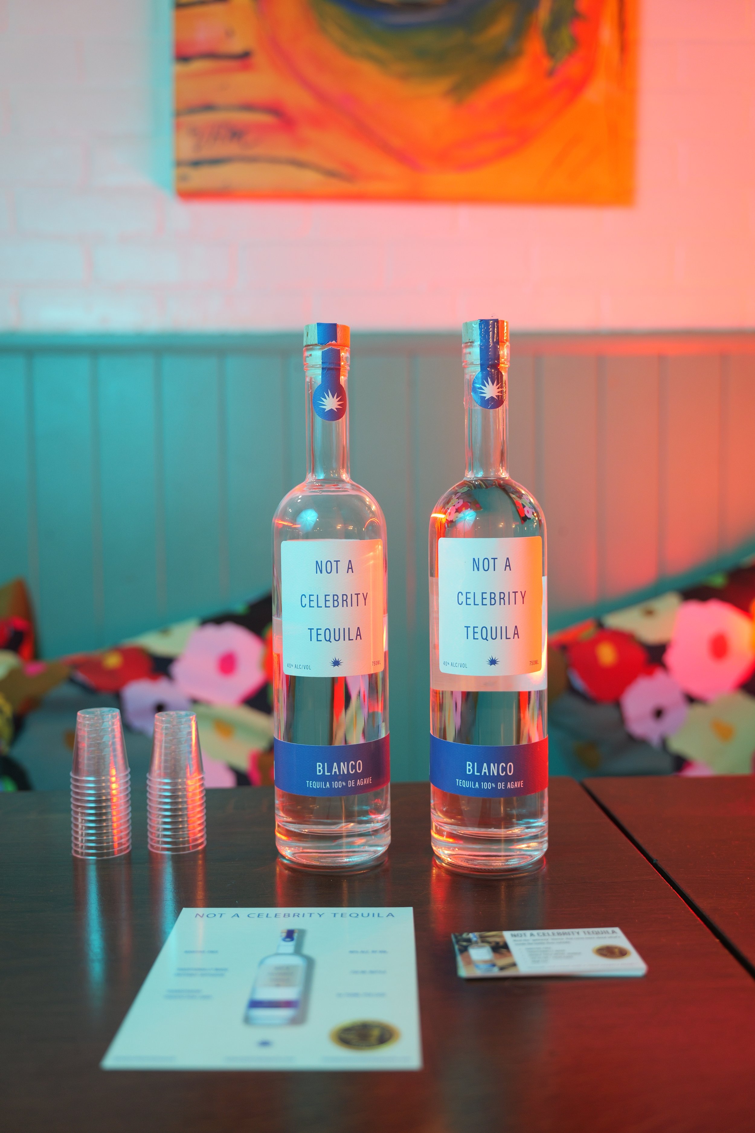 Two bottles of tequila labeled 'NOT A CELEBRITY TEQUILA' and 'BLANCO' placed on a dark wooden table, with clear plastic cups and promotional flyers in the foreground. A colorful abstract painting hangs on a white brick wall in the background, illuminated with pink and teal lighting.