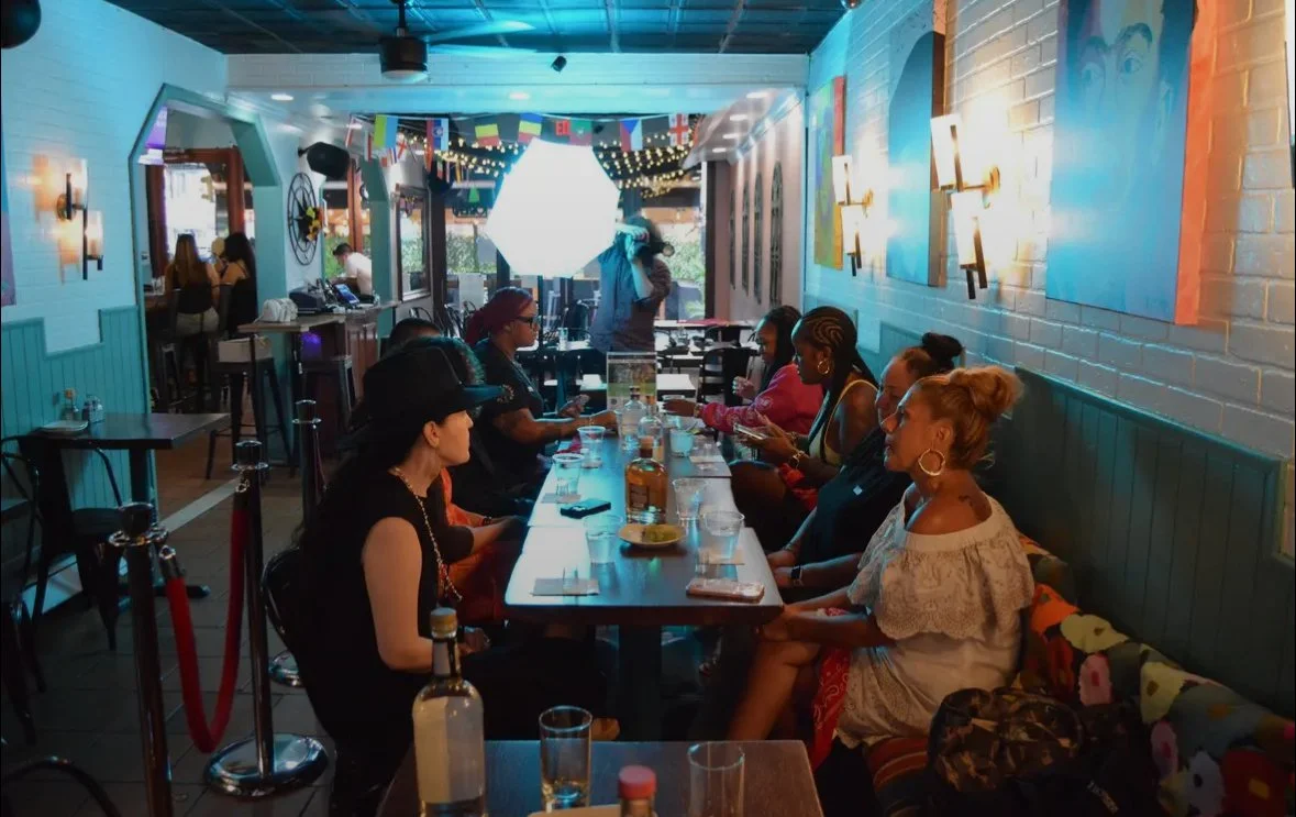 People sitting at a long table in a colorful restaurant or cafe, with string lights and flags hanging from the ceiling, some looking at their phones, with a DJ or musician performing in the background.