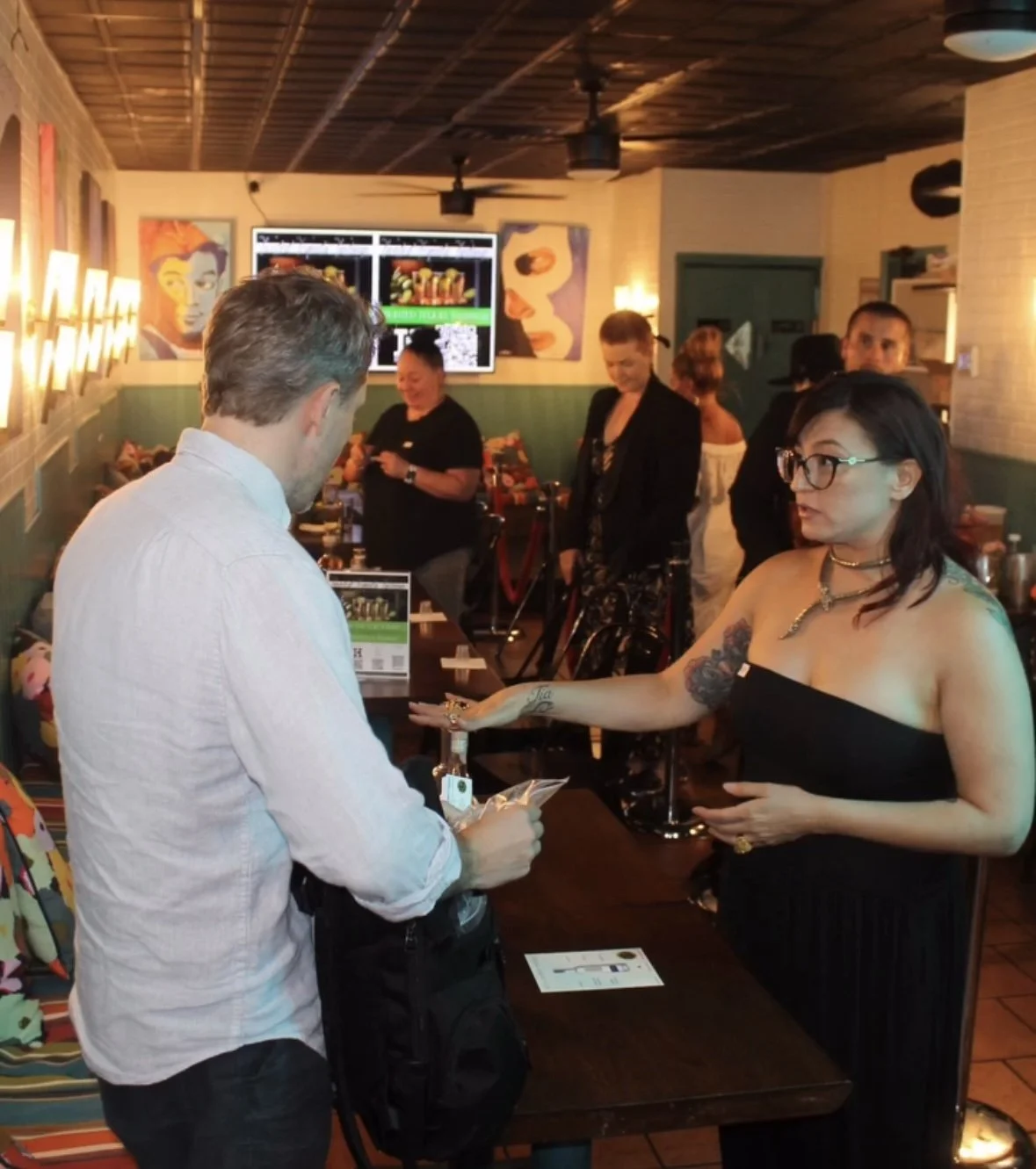 A woman with glasses, tattoos, and a black strapless dress is reaching for a bottle at a bar counter, while a man with gray hair, wearing a white shirt, faces her. Several people are in the background inside a stylish restaurant or bar with artwork on the walls.