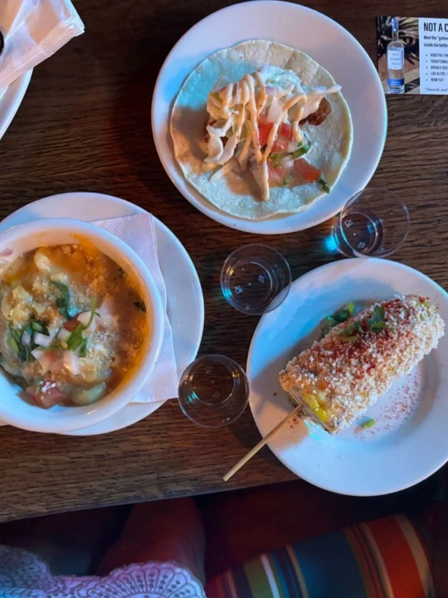 Three plates of food on a wooden table: a taco with shredded cheese, lettuce, tomato, and sour cream; a bowl of soup or stew with cheese and vegetables; and a toasted corn dog with breadcrumbs and a green garnish.