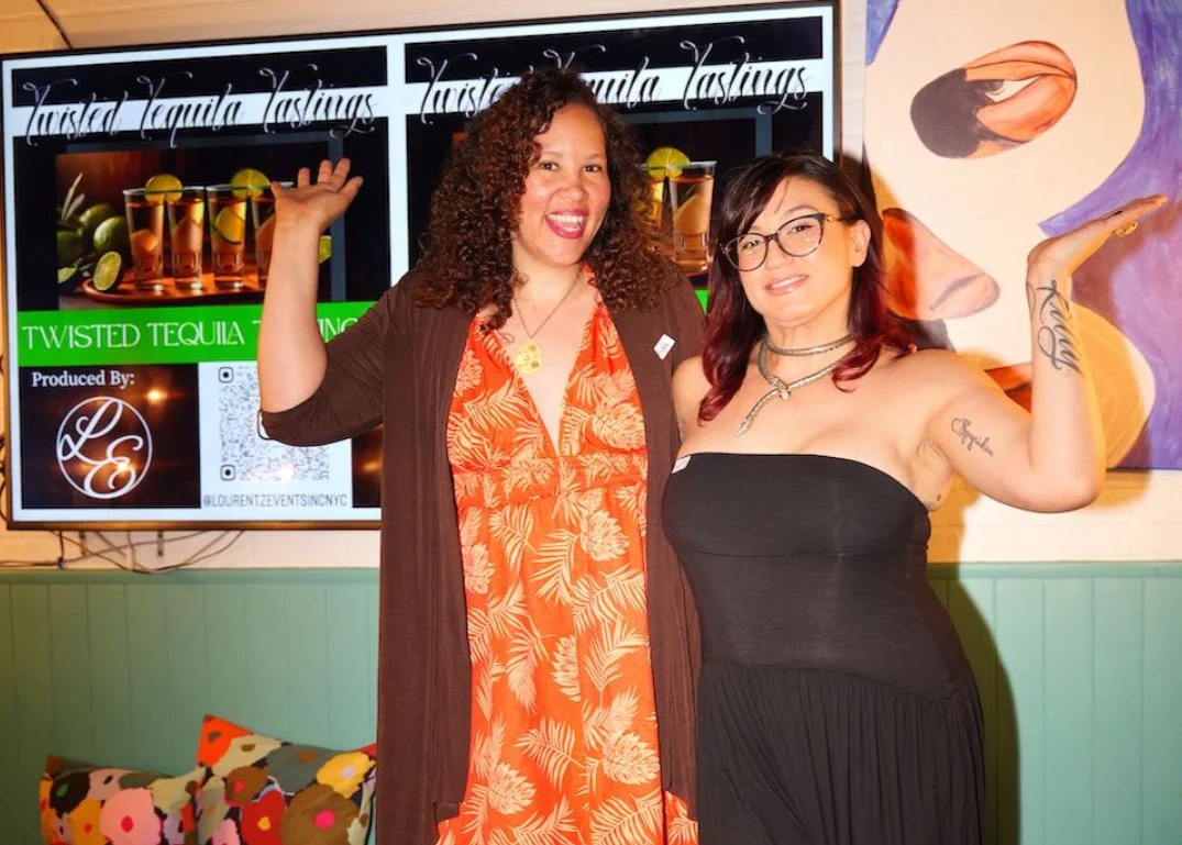 Two women standing side by side indoors in front of a large screen advertising twisted tequila tasting. The woman on the left has curly hair, wearing an orange floral dress with a brown cardigan, smiling and raising her arm. The woman on the right has shoulder-length hair, glasses, tattoos, wearing a black strapless dress, smiling, and flexing her arm.