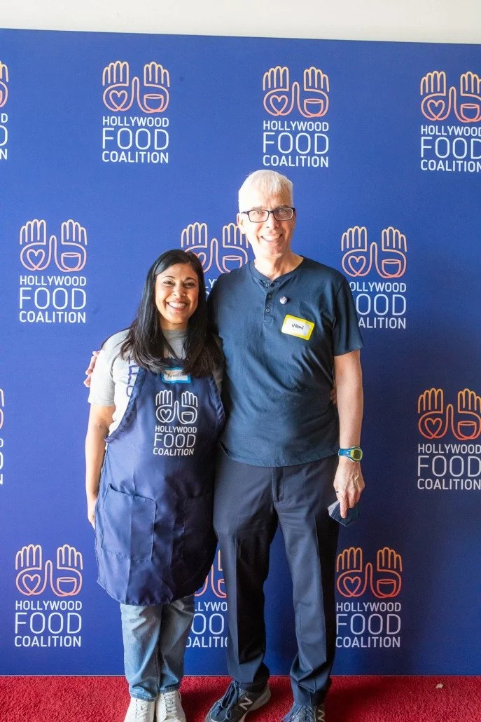 Two smiling people standing in front of a blue backdrop with the Hollywood Food Coalition logo, which features a heart and hands symbol. The woman on the left wears an apron and badge, the man on the right wears glasses and a smartwatch.