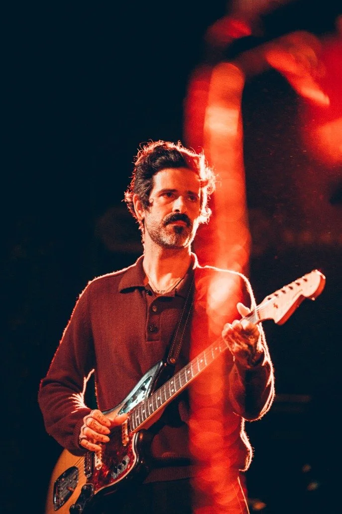 A man with dark, wavy hair and a beard playing an electric guitar on stage, illuminated by red stage lighting.