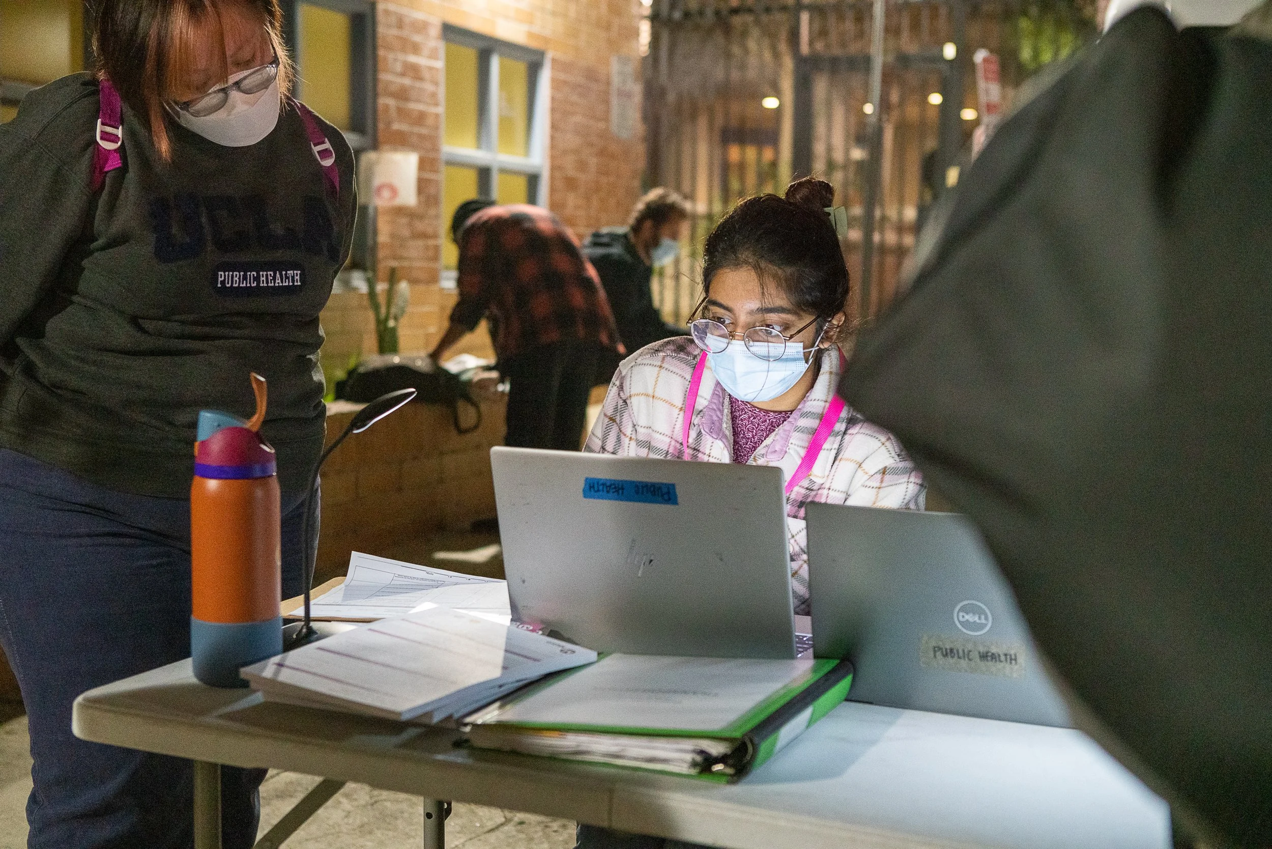 A woman wearing face mask and glasses sitting at a table with laptops, papers, and a water bottle, with two other women in the background also wearing masks. The setting appears to be an outdoor or semi-outdoor location.