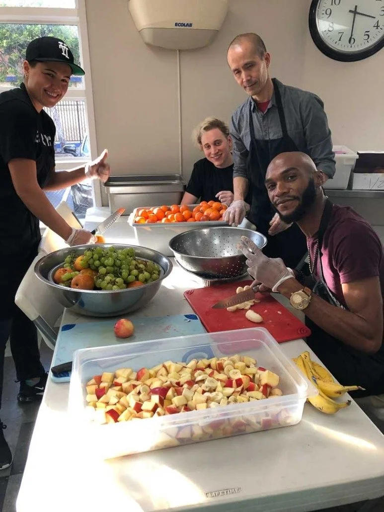Four people preparing fruit in a bright kitchen with a clock on the wall and a window showing a parking lot outside.