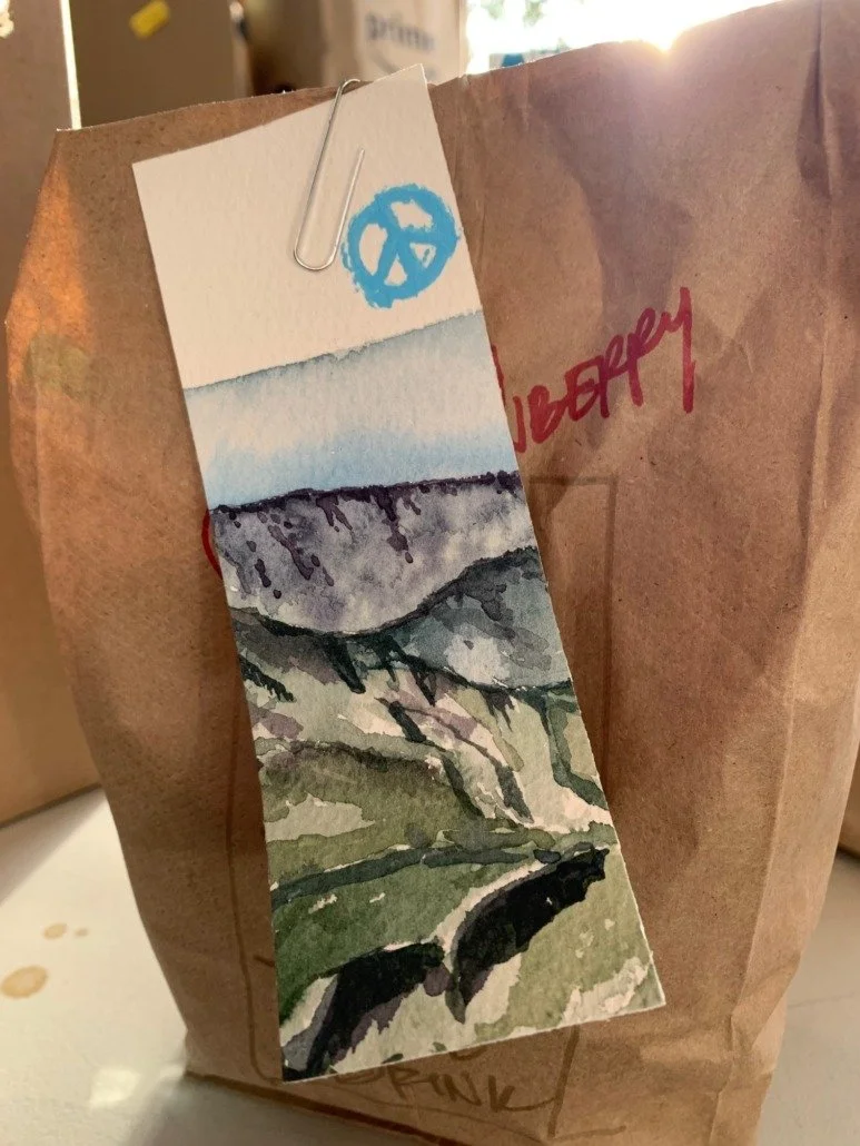 Brown paper bag with a watercolor landscape bookmark attached by a paperclip. The bookmark features mountains, trees, and a blue sky, with a blue peace symbol stamp at the top.