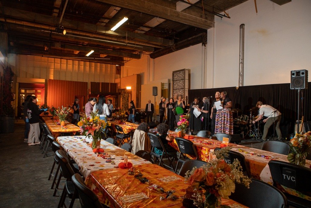 A large indoor event space with decorated tables and a stage where a choir is performing, surrounded by people and floral arrangements.