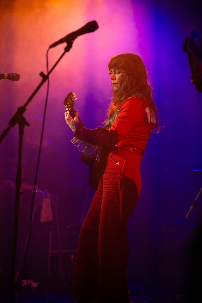 A woman with long brown hair playing an acoustic guitar on stage under colorful purple and orange lighting.