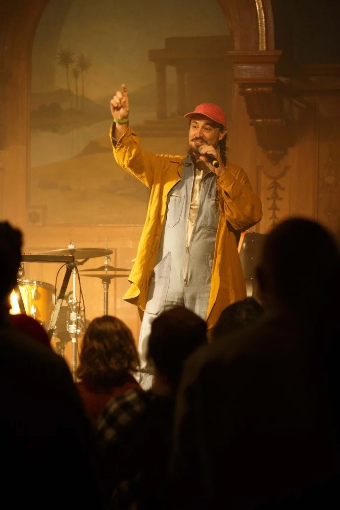 A man wearing a red cap, yellow jacket, and gray overalls speaking into a microphone on stage with a drum set behind him, audience members in front, and a scenic background in the theater.