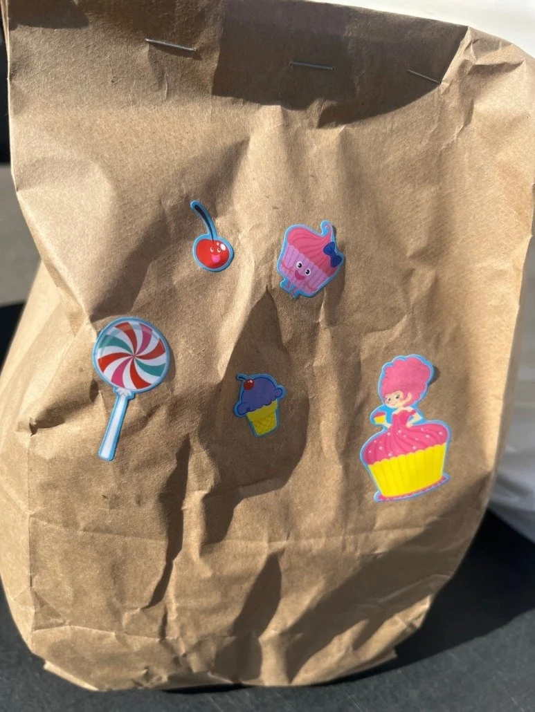 Brown paper bag decorated with colorful stickers of a cherry, a cupcake with pink frosting, a lollipop, a blue ice cream cone, and a girl in a pink cupcake dress