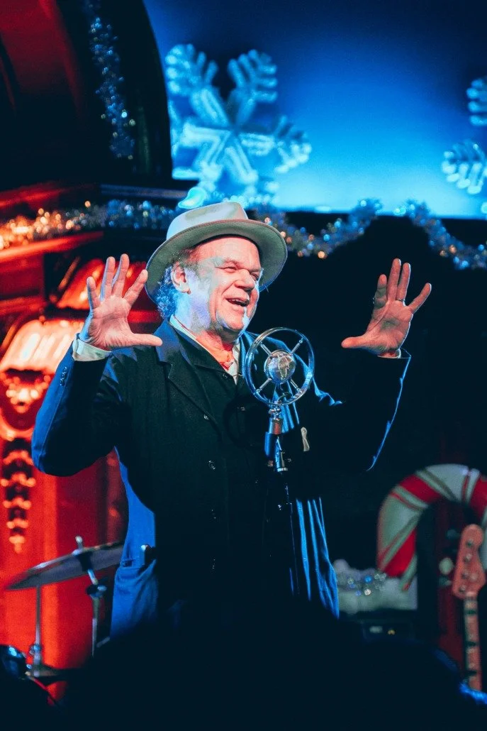 A man celebrating on stage with a vintage microphone, wearing a light-colored hat and dark clothing, decorated with Christmas-themed decorations including a snowflake background, tinsel, and a candy cane.