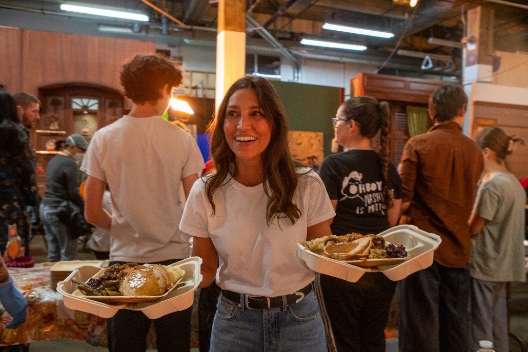 A woman holding two takeout food trays, smiling in a crowded indoor setting.