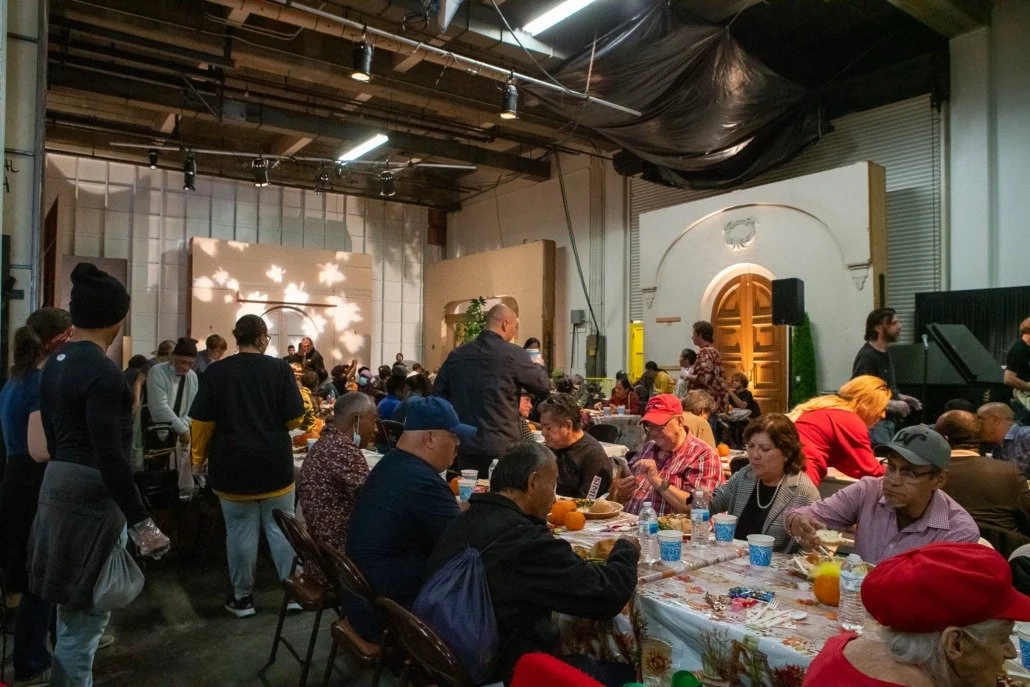 Indoor event with many people seated at tables decorated with tablecloths, pumpkins, and autumn-themed items, some standing and chatting. The background features a stage with lighting effects, a wooden door, and industrial ceiling.