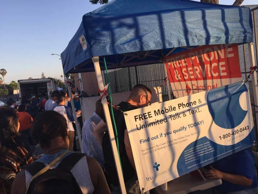 A crowd of people standing in line at a mobile phone and service booth with a blue canopy. A sign offers free mobile phones, unlimited talk and text, and includes a phone number for more information.
