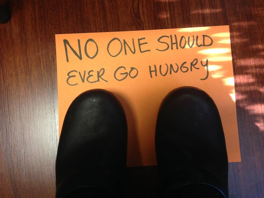 View of black shoes standing on a piece of paper with the handwritten message "No one should ever go hungry" on a wooden floor.