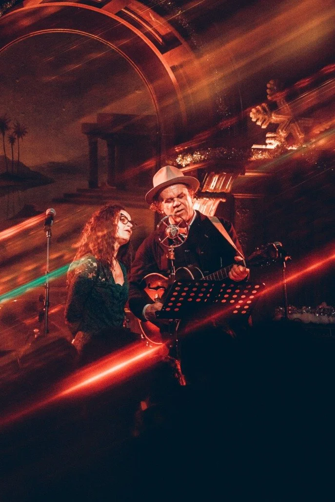 A man and woman performing music on stage with a vintage microphone and guitar, surrounded by colorful light streaks and dark background.