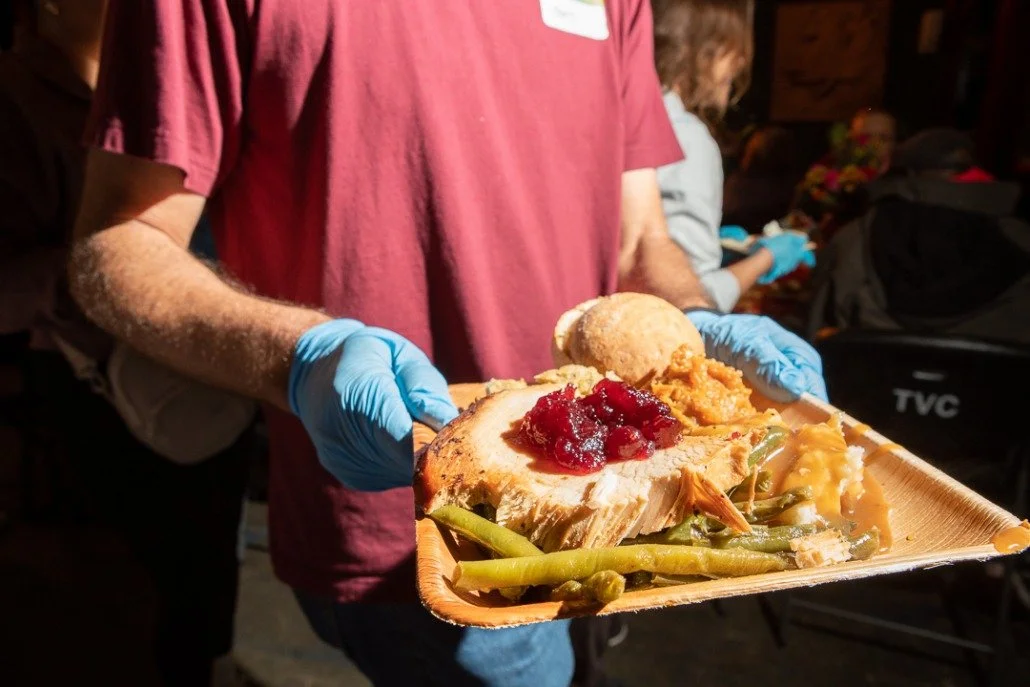 Person wearing a maroon shirt and blue gloves holding a tray with a slice of turkey, green beans, stuffing, and a bread roll.
