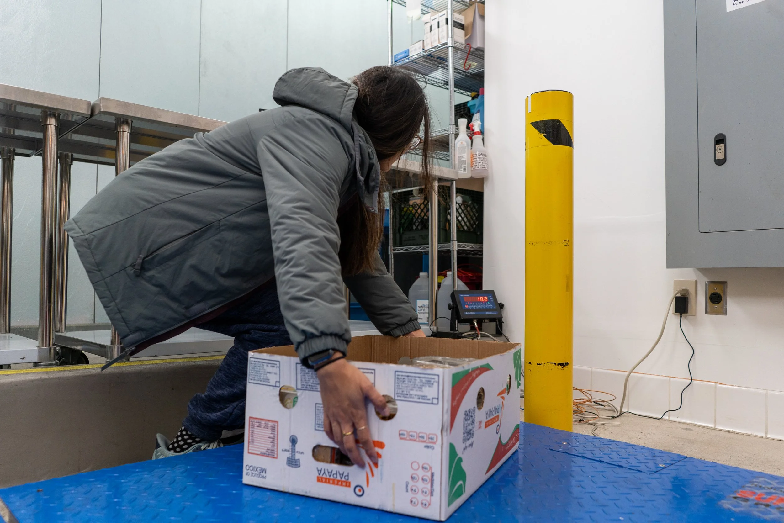 A woman bending down to pick up a cardboard box in a warehouse or storage room. The box is labeled with a produce brand, and there are shelving units with cleaning supplies in the background. The flooring is blue, and the scene includes a yellow safe