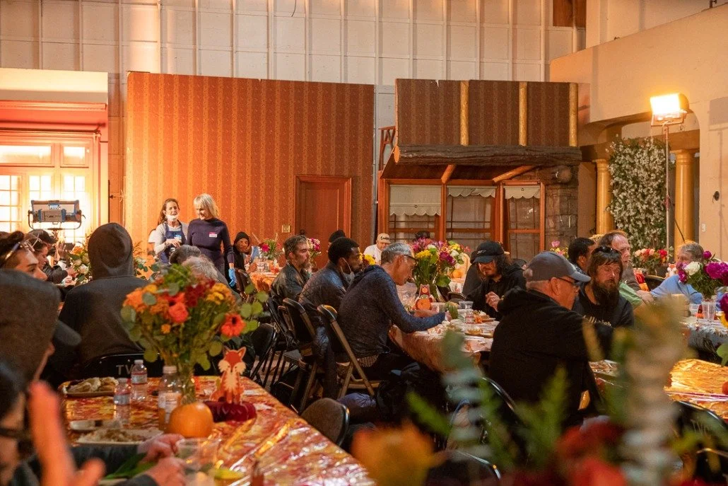 People seated at long tables with colorful flower centerpieces in a decorated indoor setting, likely a banquet or gathering.