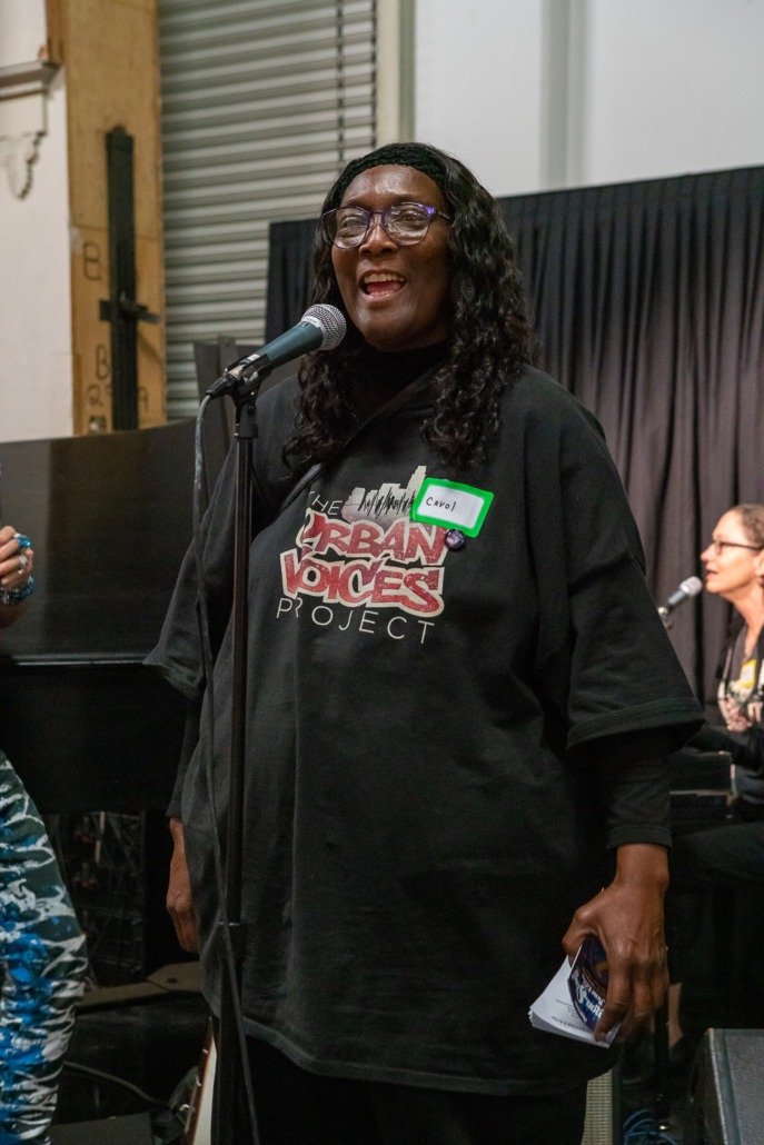 Woman with glasses speaking into a microphone at a public event, wearing a black T-shirt with the logo 'Urban Voices Project' and a green name tag that says 'Caryl'. A woman with glasses playing the piano is visible in the background.
