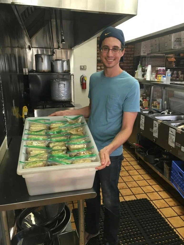 A young man wearing glasses, a blue t-shirt, and a dark blue baseball cap with a yellow Nike logo, stands in a kitchen holding a plastic container with multiple sealed bags of food. The kitchen has pots on the stove, and various cooking supplies and 