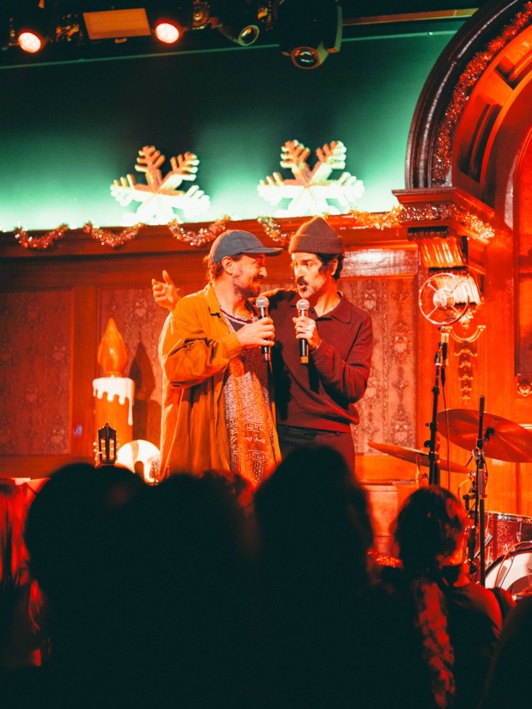 Two men with microphones on stage in a cozy venue decorated with Christmas lights and snowflake ornaments, performing or sharing a moment, audience visible in foreground.
