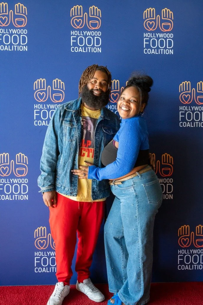 A man and woman standing in front of a blue backdrop with Hollywood Food Coalition logos. The man is wearing a denim jacket, a yellow shirt, red pants, and white sneakers. The woman is wearing a blue sweater, black top, and wide-leg jeans. They are s