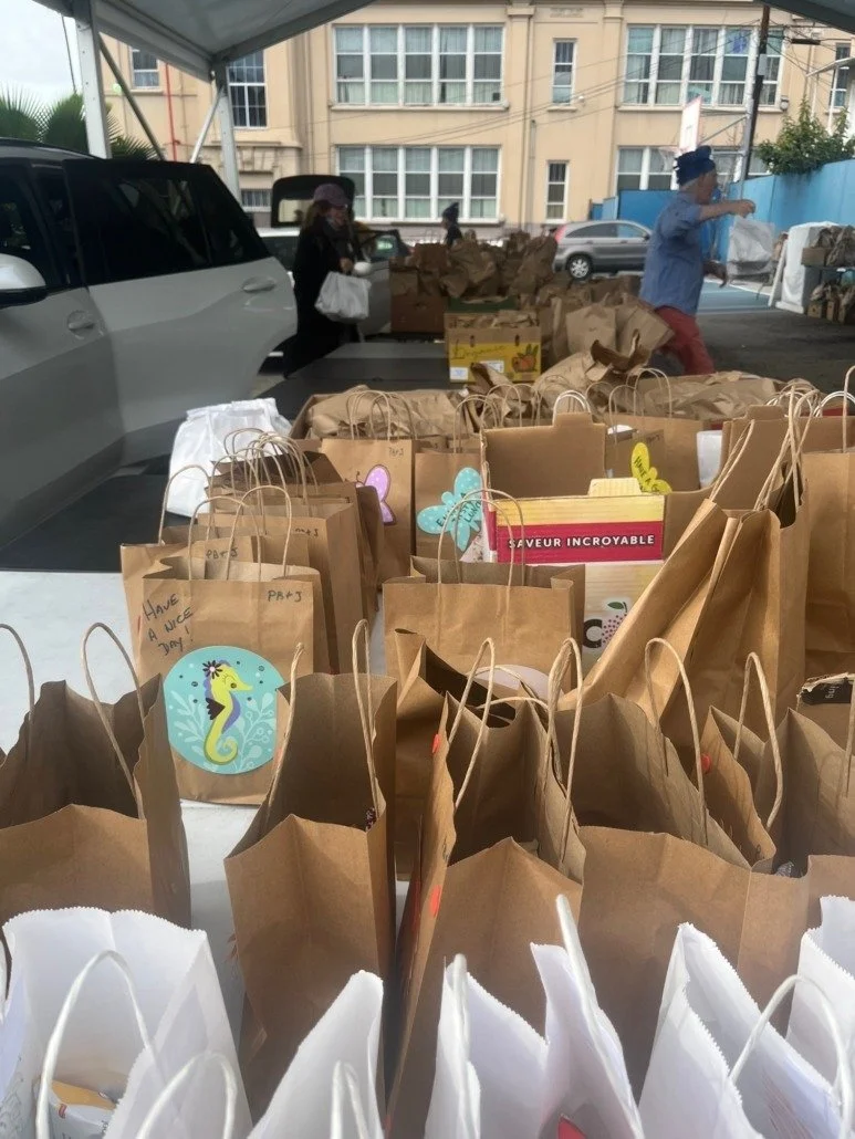 Several paper gift bags on a table, with people working behind, possibly in an outdoor setting under a tent, near parked cars and a building in the background.