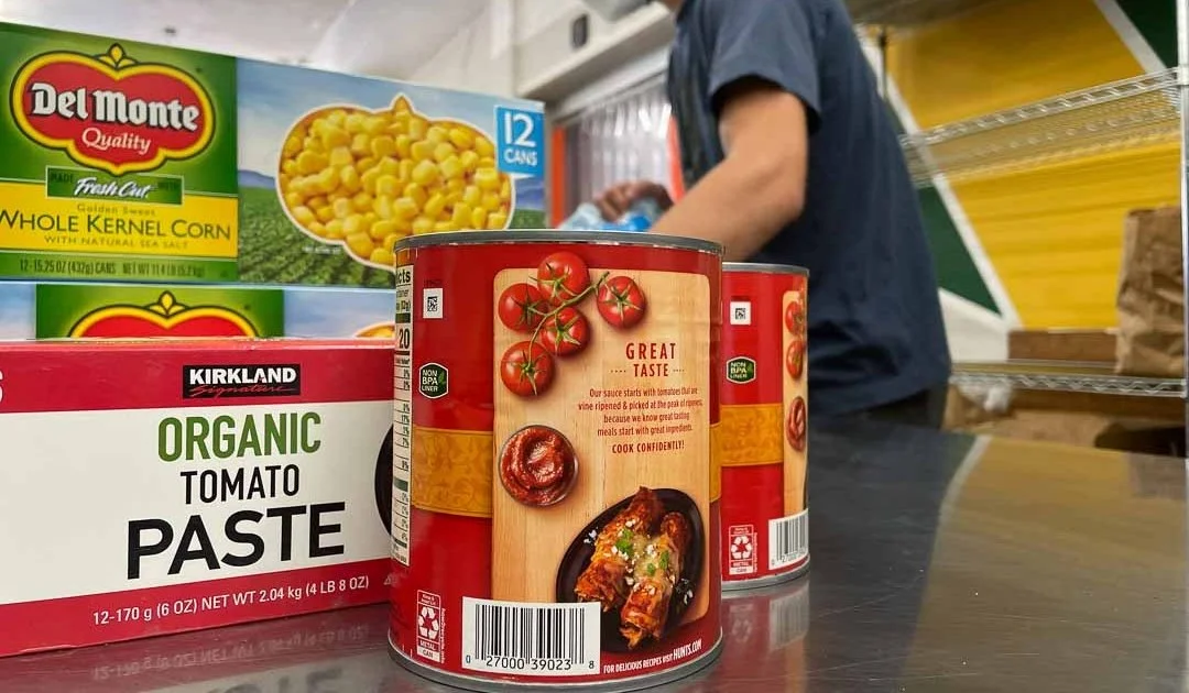Canned tomatoes, canned corn, and organic tomato paste on a store checkout counter with a person in the background.