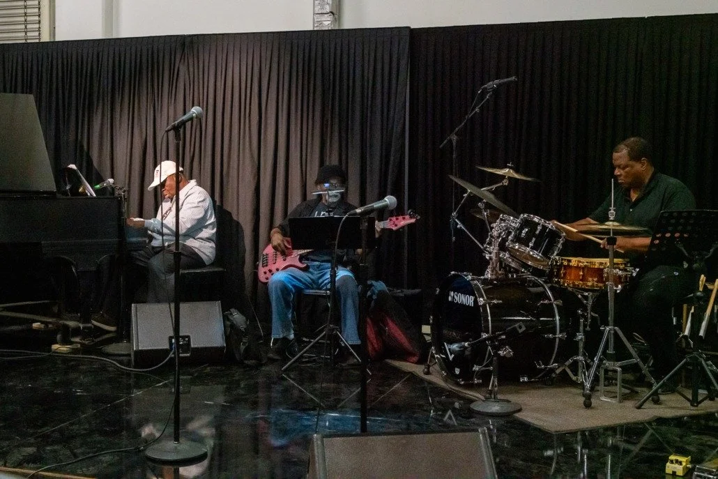 A jazz band performing on stage with a piano, an electric bass guitar, and a drum set, all set against a dark curtain backdrop.