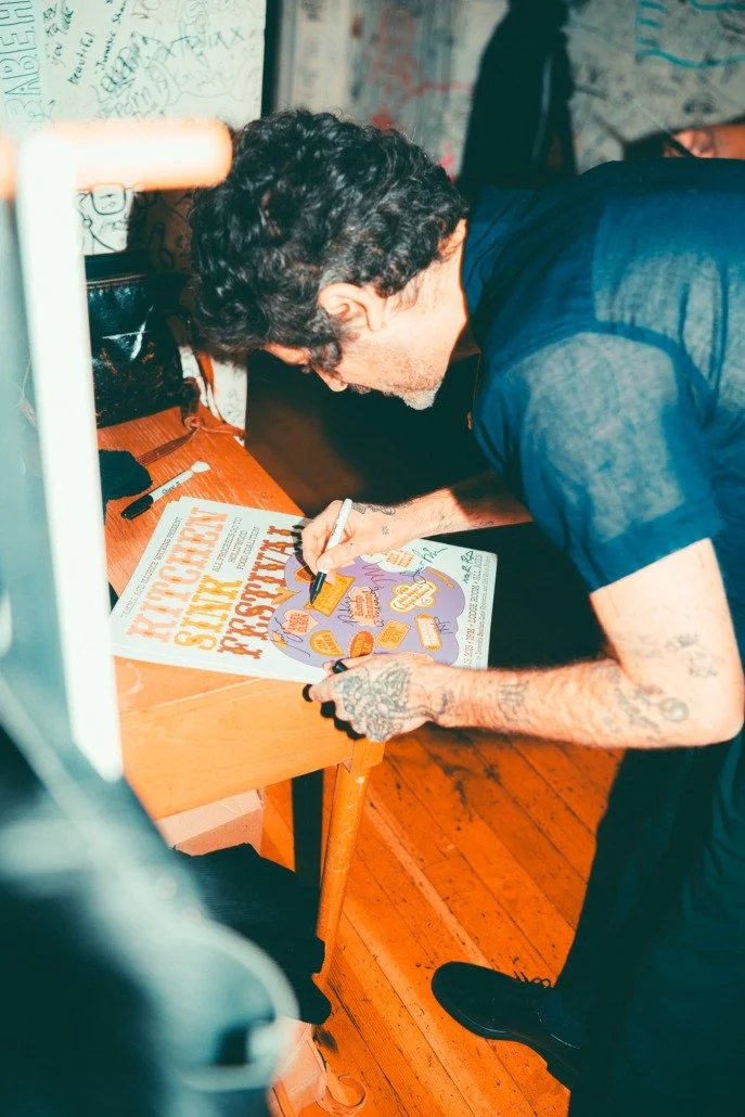 A man with tattoos on his arms signing a poster on a wooden table, with graffiti on the wall in the background.
