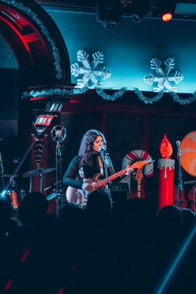 A woman with curly hair playing an electric guitar and singing into a microphone during a holiday-themed music performance. The stage is decorated with snowflakes, tinsel, a large candle prop with a flame, and Christmas candy cane decorations.