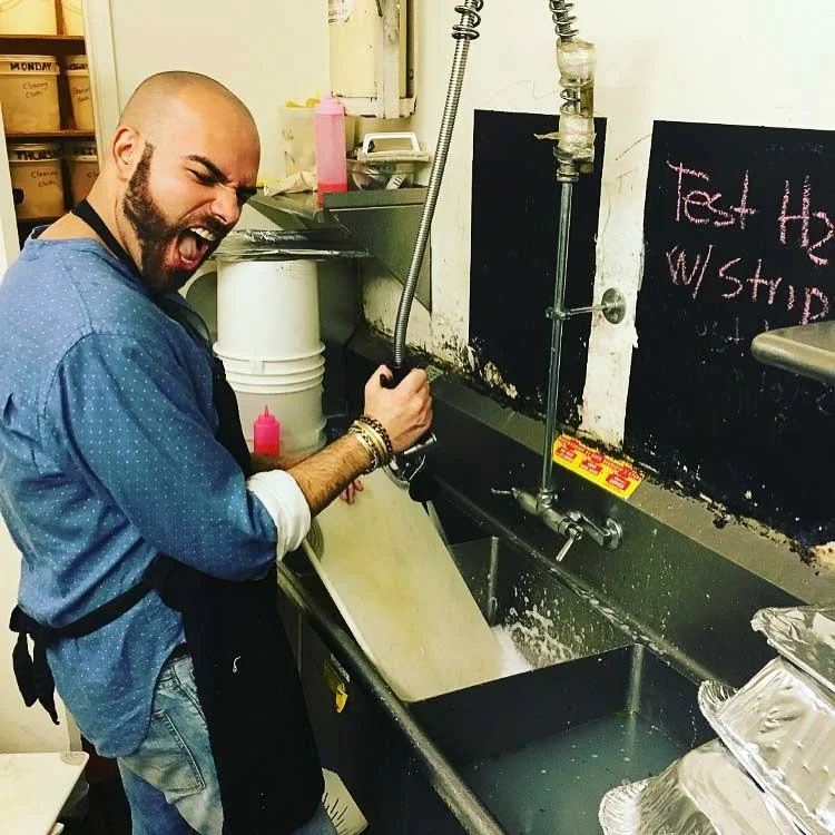 A man with a beard and a bald head is wearing a blue shirt and an apron. He is playfully making a roaring face while holding a hose over a large sink in a kitchen or café setting. Behind him are shelves and various kitchen supplies.