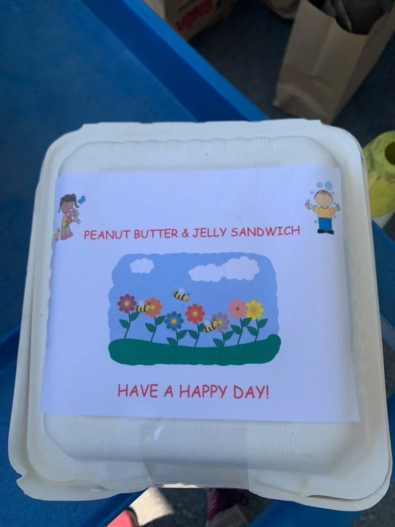 A container with a label that says "Peanut Butter & Jelly Sandwich" and "Have a Happy Day!" The label features cartoon illustrations of children and a colorful scene with flowers, bees, clouds, and a blue sky.