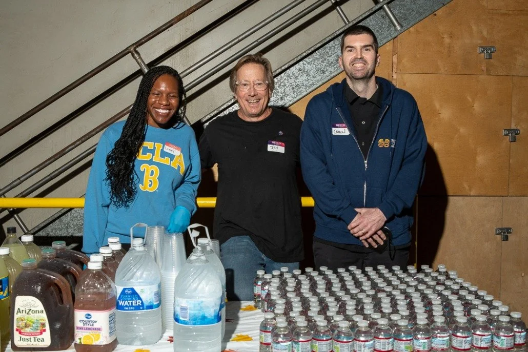 Three people standing behind a table with bottled drinks, including water, tea, and lemonade, at a community event. They are smiling with an industrial staircase in the background.