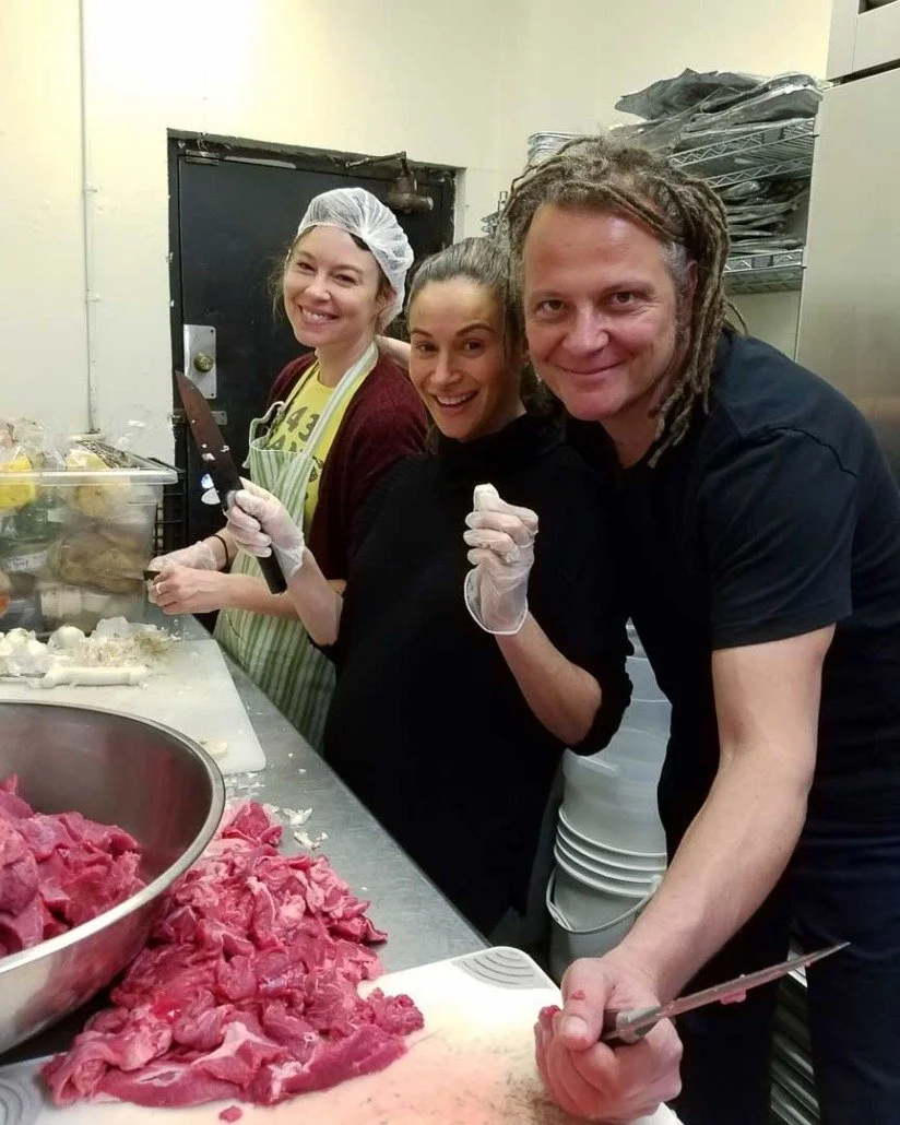 Three people preparing meat in a kitchen, smiling at the camera. One woman is wearing a hairnet, apron, and gloves, holding a knife. Another woman and a man, with dreadlocks, are also wearing gloves and holding kitchen tools, standing next to a large
