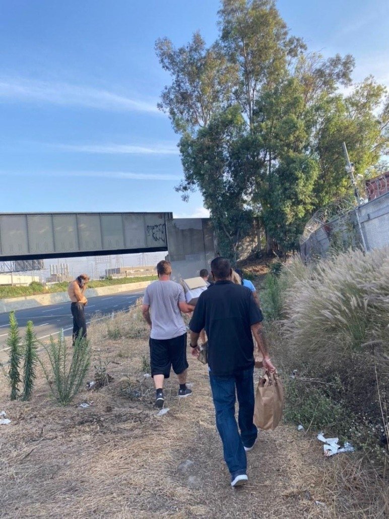 Group of people walking along a dirt path beside a highway with an overpass, trees, and dry grass on the side.
