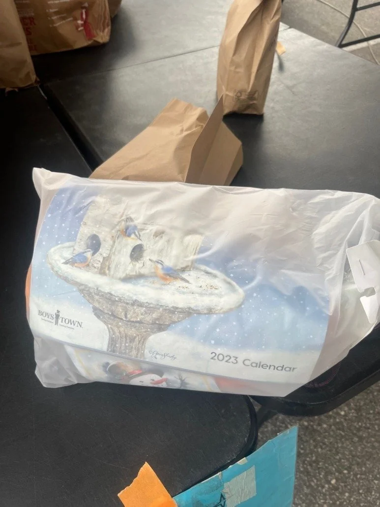 2023 calendar with a winter-themed cover featuring ducks around a birdbath, placed on a black surface among paper bags.