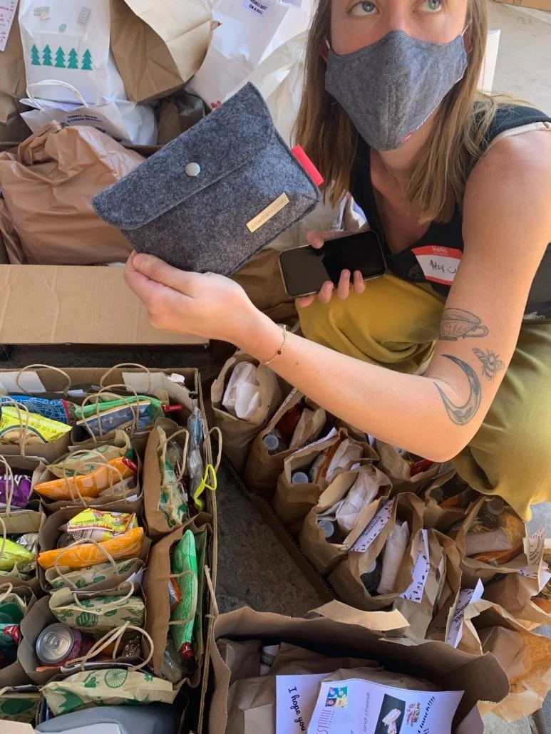 Woman in gray face mask holding a gray felt pouch and a smartphone, surrounded by paper bags filled with snacks and supplies.