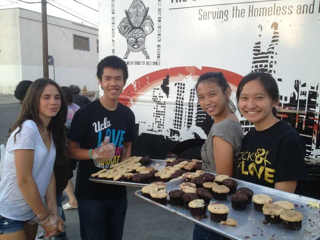 Four young people standing in front of a food truck with trays of cookies, smiling, at an outdoor event. Two women and two men, with the woman on the far left and the woman second from the right wearing black T-shirts with yellow text.