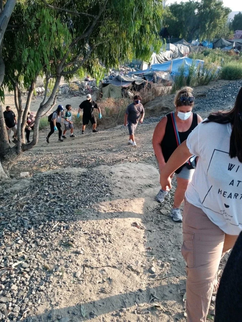 A group of people, including children and adults, are walking up a dirt and gravel hill outdoors, with some wearing masks. In the background, there are makeshift tents and shelters among trees.