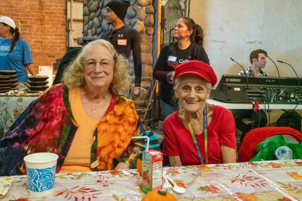 Two elderly women smiling at a table during a gathering, one wearing a red hat and the other with curly blonde hair and colorful clothing. Other people and a keyboard are visible in the background.