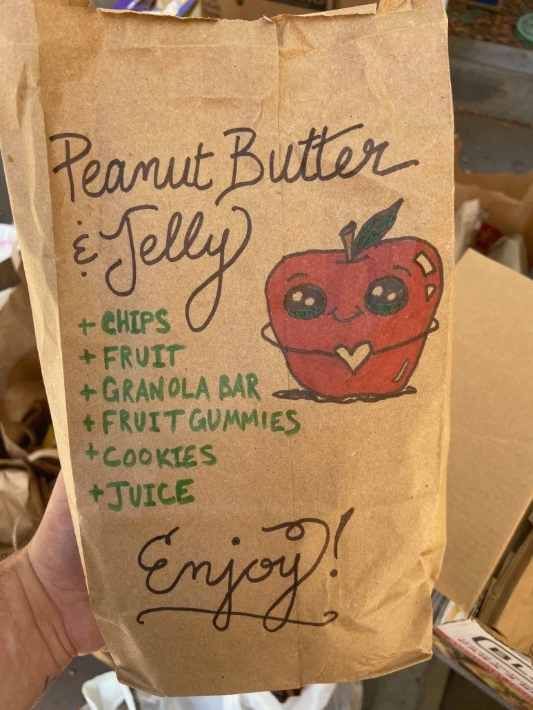 Paper bag with colorful handwritten list of treats and a cute drawn apple with face, arms, and a heart-shaped belly button. The list includes peanut butter & jelly, chips, fruit, granola bar, fruit gummies, cookies, and juice, with a message saying "