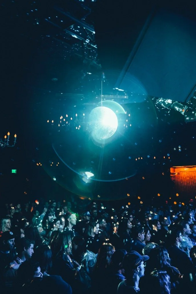 Audience watching a concert or event with a large, bright, glowing object overhead, possibly a light or installation, in a dark venue.