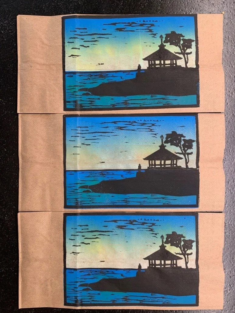 Three identical landscape art prints on brown paper backgrounds, each depicting a waterside scene with a house, trees, and a person, set against a colorful sky.