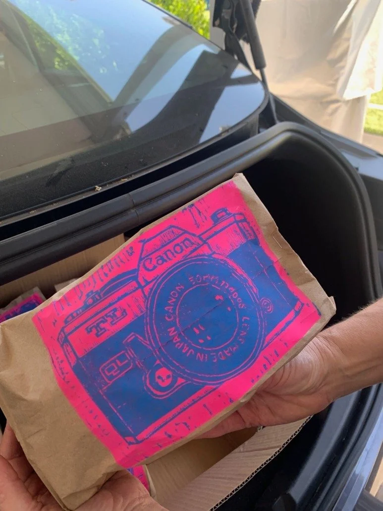 Person holding a brown paper bag with a pink and blue illustration of a Canon camera on it, placed inside a car's trunk.