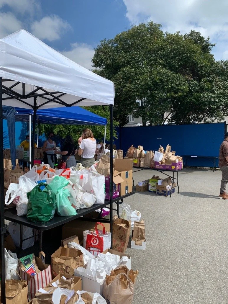 Outdoor charity event with tables and tents filled with paper shopping bags, some with visible logos, on a paved surface. Several people are in line or shopping, with a large tree and blue sky in the background.