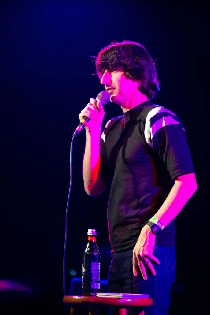 A young man with dark, curly hair stands on stage holding a microphone, wearing a black sports shirt with white accents. There is a bottle of soda on a small table in front of him, and he is illuminated by purple stage lighting.