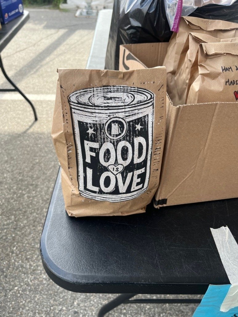 A paper bag with a black and white illustration of a soda can that reads 'Food is Love' on a black table, with other paper bags partially visible.